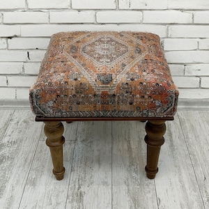 Pode incluir: Um pequeno otomano quadrado com um topo de tecido estampado de cores e pernas de madeira. O tecido apresenta um design geométrico em tons de laranja, vermelho e azul.