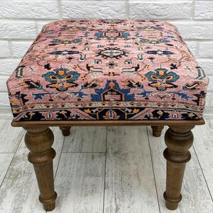 May include: A pink and blue patterned ottoman with wooden legs. The ottoman has a traditional rug design.