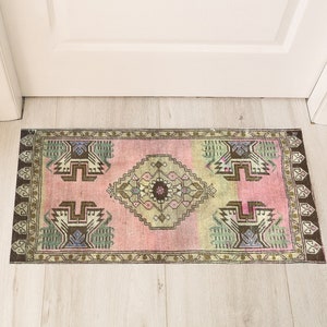 以下が含まれることがあります： 幾何学模様のピンクとグリーンの敷物。敷物は白いドアの前にある明るい木製の床に置かれています。