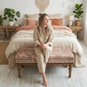 May include: A bedroom with a bed dressed in neutral-toned bedding and cushions. A patterned bench is at the foot of the bed. The room is decorated with plants and artwork, creating a relaxed and natural atmosphere. The room has a natural, relaxed aesthetic.