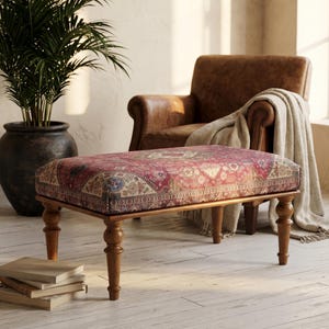 May include: A rectangular ottoman featuring a red, blue, and beige patterned fabric upholstery with a repeating geometric design. The ottoman is supported by four turned wooden legs in a medium brown finish, providing a stable base for the seat.