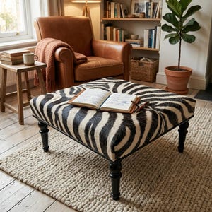 May include: A square ottoman upholstered in a black and white zebra print fabric, featuring four dark turned wooden legs. An open notebook with handwritten text and a brown pen are resting on the top surface of the ottoman.