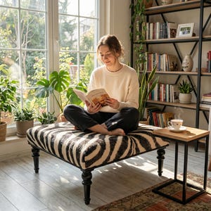 May include: A square ottoman featuring a black and white zebra print fabric upholstery, supported by four dark-toned, turned wooden legs. The piece is positioned in a bright room, serving as a seat for reading, with a simple, clean-lined design.