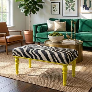 May include: A living room scene featuring a zebra-print bench with yellow legs, a green velvet sofa, and a brown leather armchair. Botanical prints adorn the wall above the sofa. A large potted plant adds a natural element to the room.