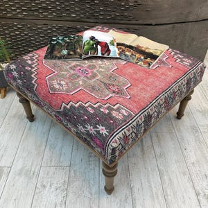 May include: A square ottoman with a red and gray patterned rug top. The rug design features a central medallion and border details. The ottoman has four wooden legs and is topped with open magazines.
