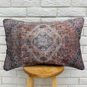 Könnte beinhalten: Rechteckiges Zierkissen mit Vintage-Teppichmuster. Das Kissen zeigt ein detailliertes Muster in Rost-, Braun- und Grautönen mit einem zentralen rautenförmigen Motiv. Das Kissen liegt auf einem kleinen Holzschemel vor einer weißen Backsteinmauer.