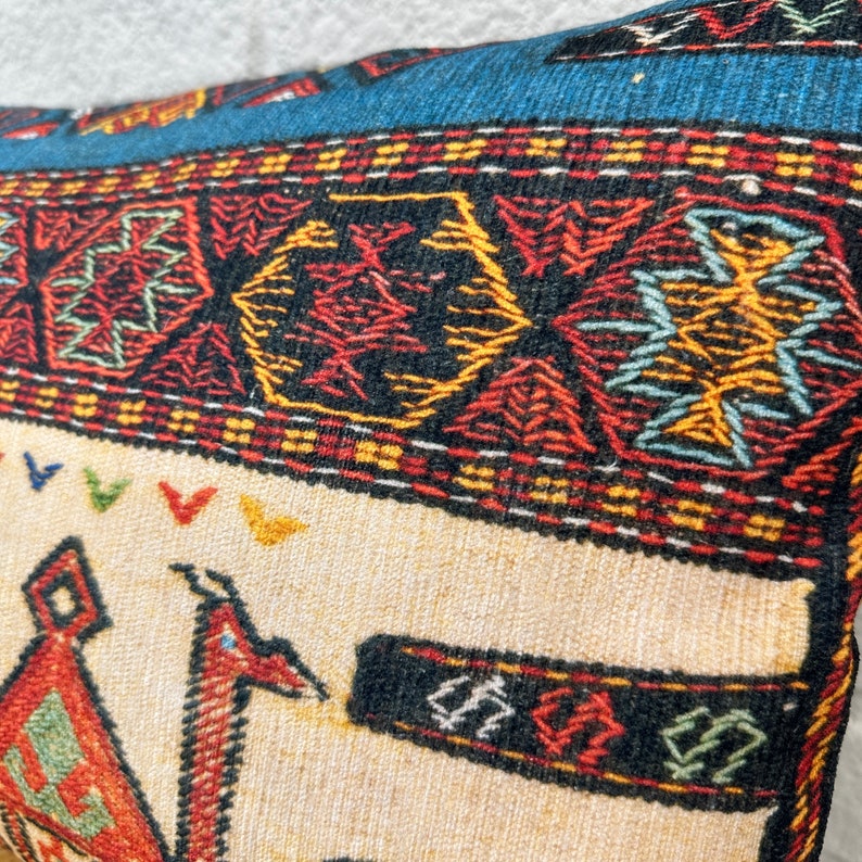 May include: Close-up of a decorative pillow with a detailed, colorful pattern. The pillow features a cream-colored central panel with a red and black design. The top border is blue with a geometric pattern.