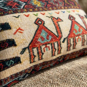 May include: Close-up of a decorative pillow with a camel design. The pillow features a beige background with a repeating pattern of red, orange, and teal camels. The edges are trimmed with a patterned border in shades of red, black, and gold.