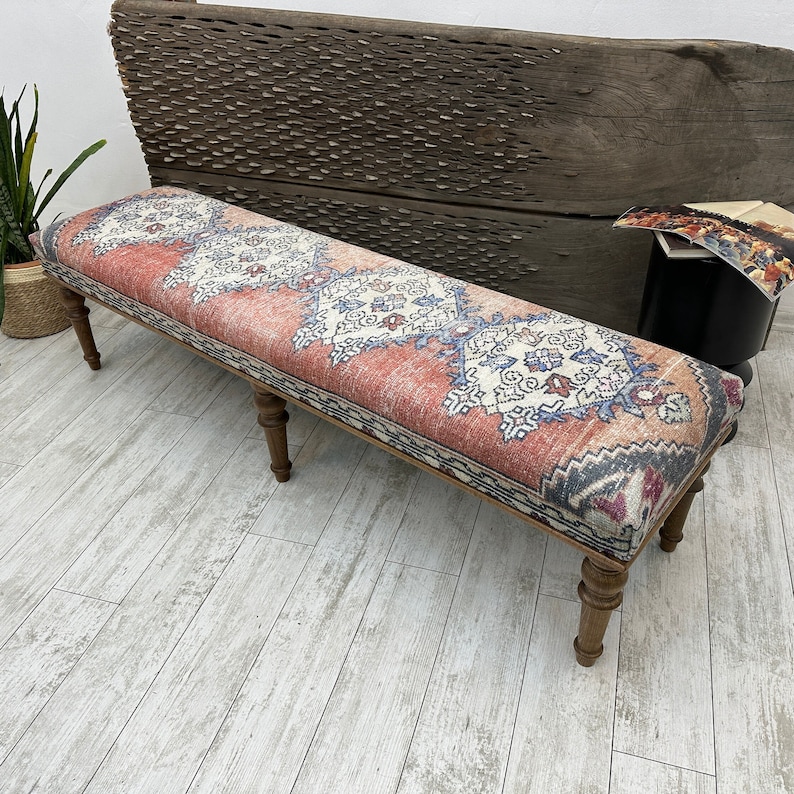 May include: A long, rectangular bench with a red, white, and blue patterned fabric top. The bench has four wooden legs and is sitting on a light wood floor.