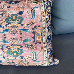 May include: A decorative pillow with a Persian rug design. The pillow features a pink background with a detailed pattern in shades of blue, orange, and cream. The border is a dark blue with an intricate design. The pillow is resting on a dark gray surface.