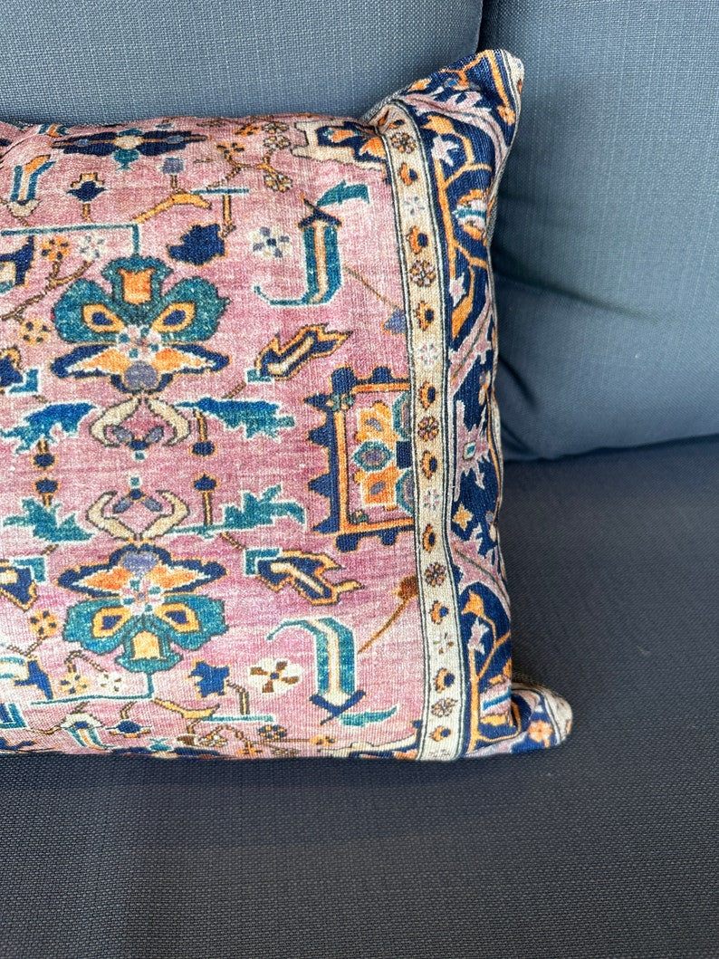 May include: A decorative pillow with a Persian rug design. The pillow features a pink background with a detailed pattern in shades of blue, orange, and cream. The border is a dark blue with an intricate design. The pillow is resting on a dark gray surface.
