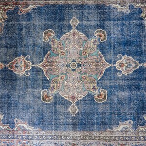 Op de afbeelding: Een vintage blauw tapijt met een vervaagd bloemenpatroon in het midden. Het tapijt heeft een rand met een herhalend patroon.