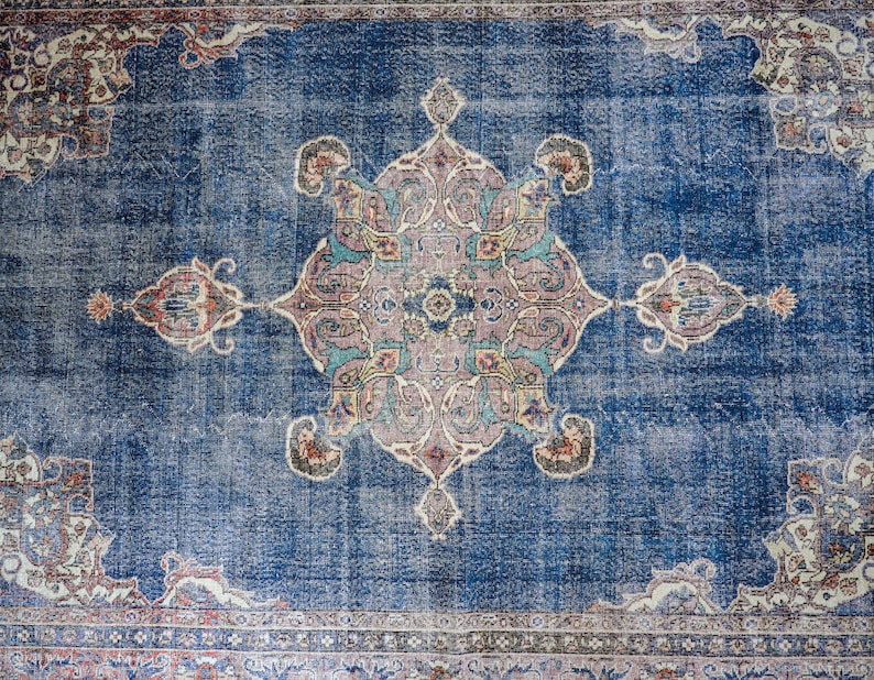 Op de afbeelding: Een vintage blauw tapijt met een vervaagd bloemenpatroon in het midden. Het tapijt heeft een rand met een herhalend patroon.