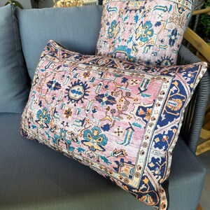 May include: Two decorative throw pillows with a vintage rug design. The pillows feature a pink and blue floral pattern with a navy blue border. They are placed on a gray outdoor sofa, creating a cozy and stylish look.