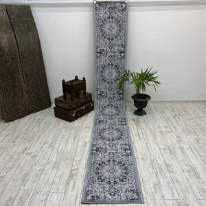 May include: A gray and white patterned runner rug with a traditional design. The rug is hanging on a wall in a room with white walls and wooden floors.