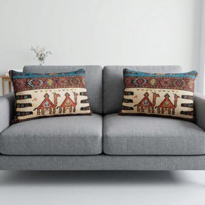 May include: Two decorative throw pillows with a camel design rest on a gray sofa. The pillows feature a cream-colored background with a camel motif in shades of brown, red, and blue. The top border is a geometric pattern with a teal accent.