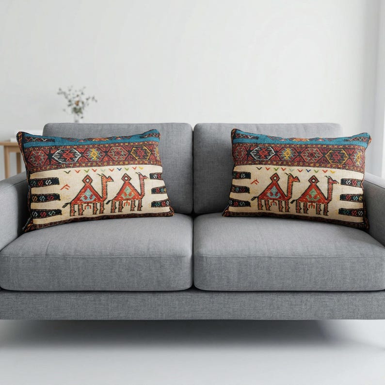 May include: Two decorative throw pillows with a camel design rest on a gray sofa. The pillows feature a cream-colored background with a camel motif in shades of brown, red, and blue. The top border is a geometric pattern with a teal accent.