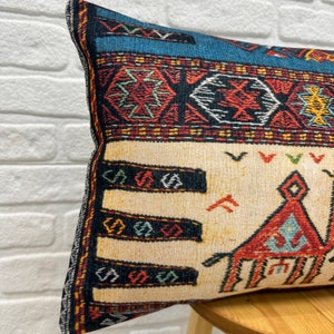 May include: Rectangular decorative pillow with a woven design. The pillow features a cream-colored background with a geometric pattern in red, blue, and black. The edges are trimmed with a patterned border.