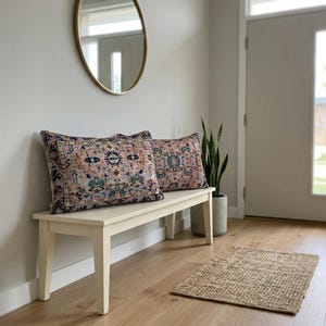 May include: Two decorative throw pillows with a vintage-inspired design rest on a cream-colored bench. The pillows feature a pink background with blue, green, and gold patterns. A woven rug and a potted plant complete the entryway decor.
