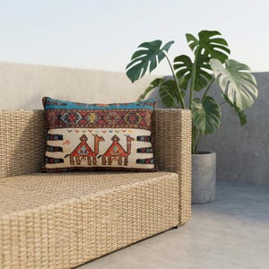 May include: Rectangular decorative pillow with a camel design. The pillow features a cream-colored background with two camels in red and orange, and a blue and patterned border. The pillow is placed on a woven wicker sofa.