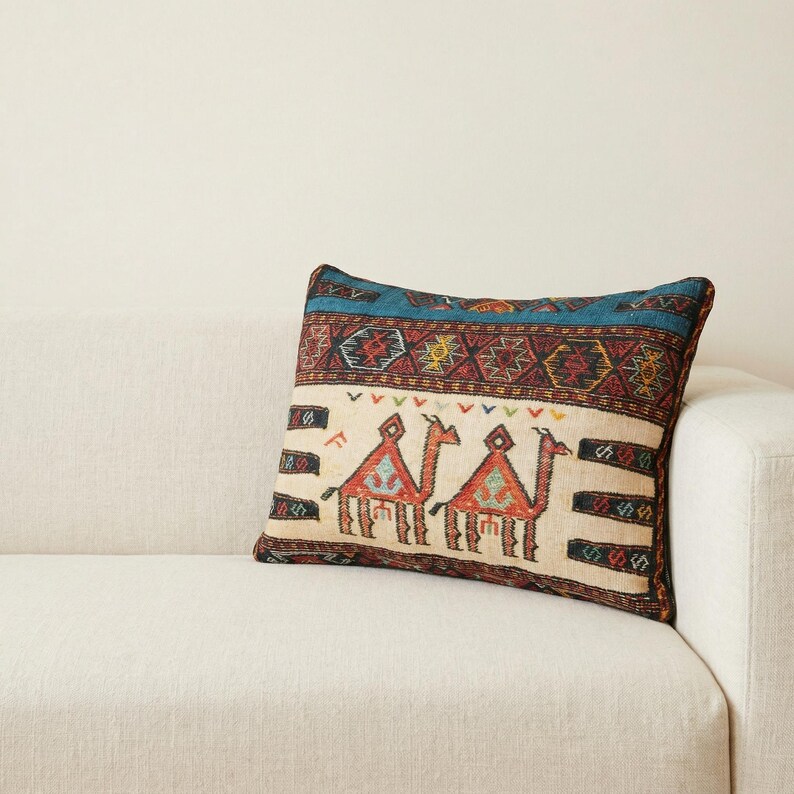 May include: Rectangular decorative pillow with a beige background and a colorful geometric pattern. The pillow features two camels in red and orange, with a blue and brown border. The pillow is resting on a white sofa.