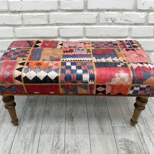 Puede incluir: Un otomano colorido y estampado con patas de madera. El otomano está cubierto con un mosaico de tela roja, naranja, azul y negra.