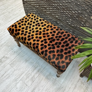 May include: A rectangular bench with a leopard print pattern in shades of brown and black. The bench has four wooden legs and is placed on a light-colored wooden floor. The bench is a decorative furniture piece.