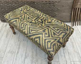 May include: A wooden bench with a geometric patterned fabric seat in shades of gold and black. The bench has turned legs and is photographed on a wood floor.
