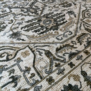 May include: A close-up of a beige rug with a detailed black and gray patterned design. The rug has a woven texture and a traditional style.