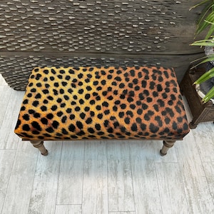 May include: A rectangular ottoman with a leopard print pattern in shades of orange, yellow, and black. The ottoman has a wooden frame and four short, light-colored legs. The overall design is reminiscent of animal print.