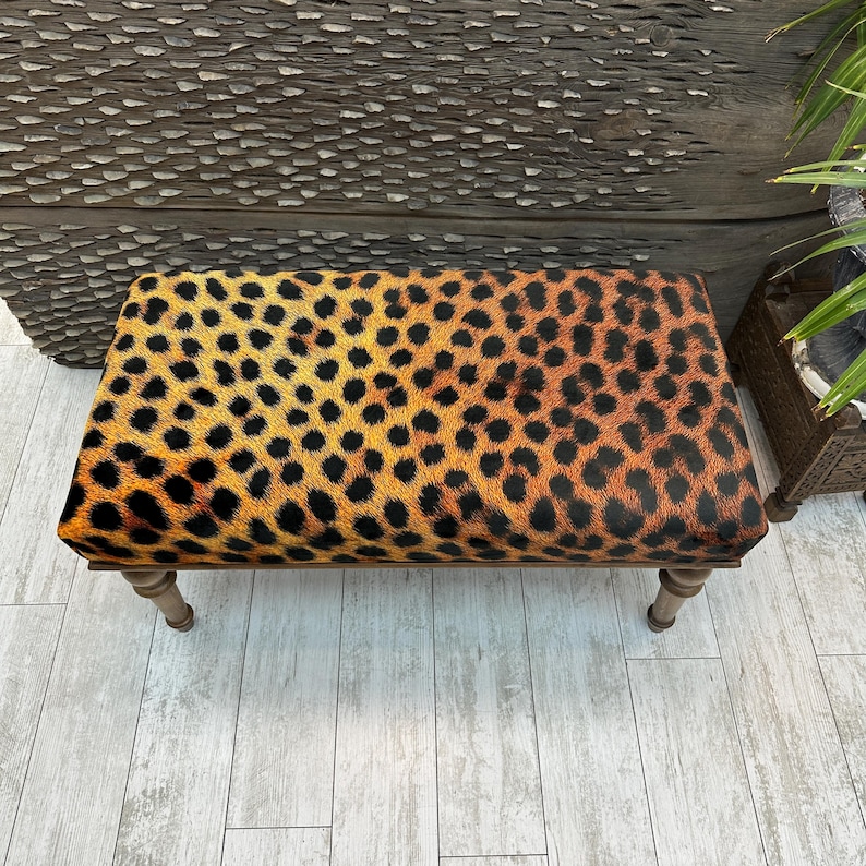 May include: A rectangular ottoman with a leopard print pattern in shades of orange, yellow, and black. The ottoman has a wooden frame and four short, light-colored legs. The overall design is reminiscent of animal print.