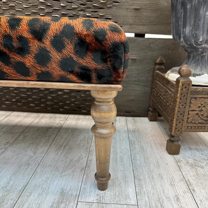 May include: A small, upholstered bench with a leopard print pattern in black and orange. The bench has a wooden frame and legs. The legs are turned and have a light brown finish. The bench is likely used for seating or as a decorative accent piece.