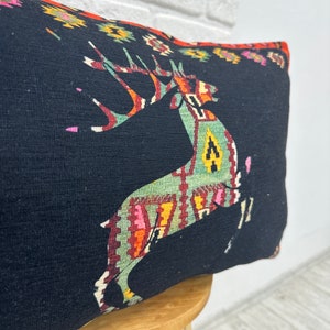 May include: Black cushion with a colourful geometric pattern featuring a stylised deer. The cushion is made of a textured fabric and has a fringed edge.