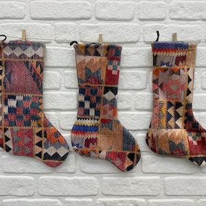 Puede incluir: Tres calcetines de Navidad coloridos con un patrón geométrico, colgados en una pared de ladrillo blanco.