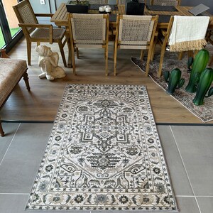 May include: A beige and brown patterned rug with a geometric design. The rug is laid out on a light gray tiled floor in a room with wooden furniture.