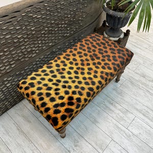 May include: A rectangular bench with a leopard print seat cushion. The cushion is a mix of yellow, orange, and black spots. The legs are made of wood and have a light brown finish. The bench is a decorative furniture piece.