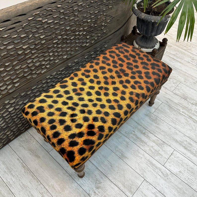 May include: A rectangular bench with a leopard print seat cushion. The cushion is a mix of yellow, orange, and black spots. The legs are made of wood and have a light brown finish. The bench is a decorative furniture piece.