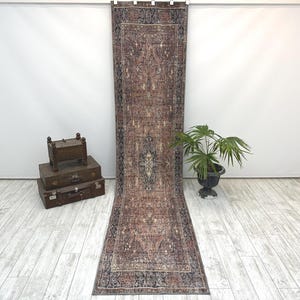 May include: A long, rectangular runner rug featuring a faded, intricate pattern in shades of dusty rose, charcoal grey, and cream. The rug has a central medallion design surrounded by a detailed border, displayed vertically against a plain wall.
