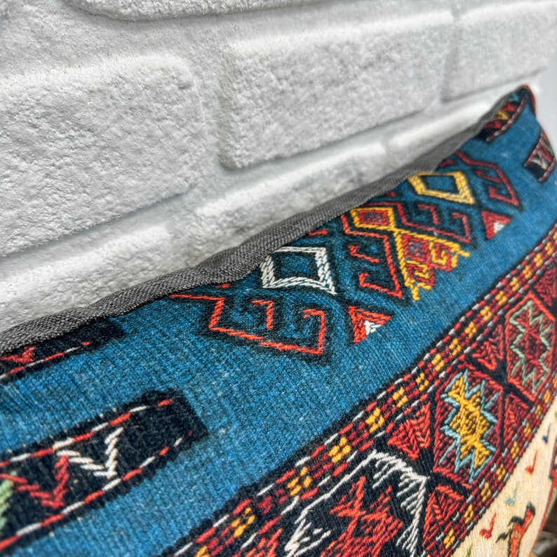 May include: Close-up of a decorative pillow with a blue background and intricate geometric patterns in red, yellow, and white. The pillow has a gray border and a textured appearance, suggesting a woven fabric.