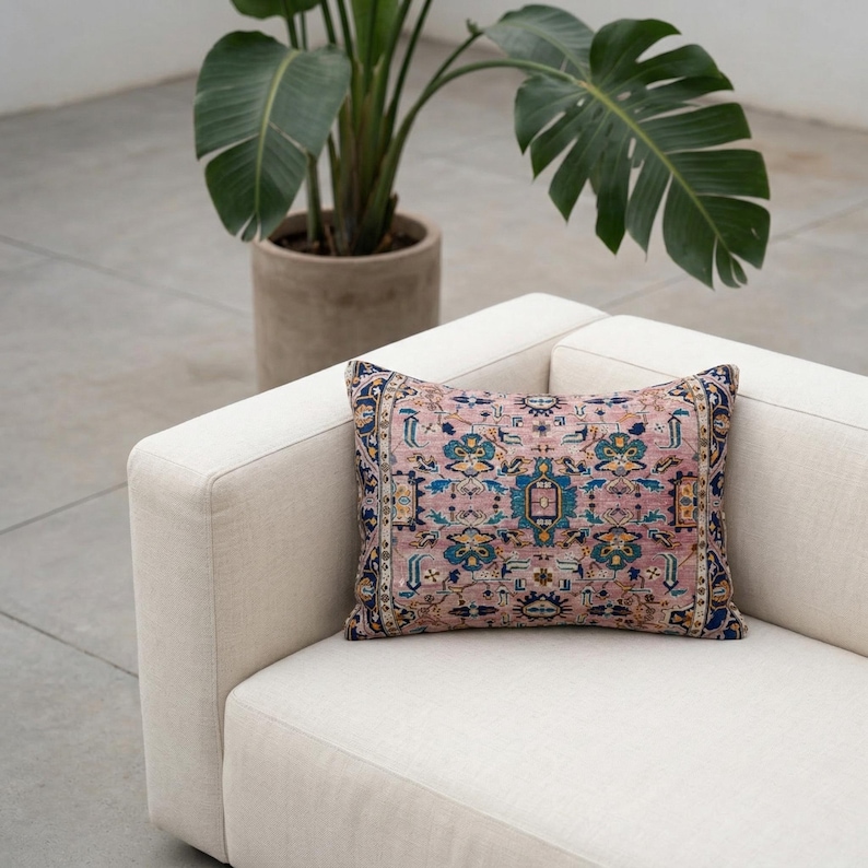May include: A decorative rectangular pillow with a vintage rug design in shades of pink, blue, and yellow. The pillow is placed on a cream-colored sofa, with a potted plant in the background.