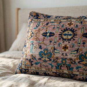 May include: A decorative pillow with a vintage rug design. The pillow features a dusty rose background with intricate patterns in navy blue, teal, and gold. The pillow is rectangular and placed on a bed with neutral-colored linens.