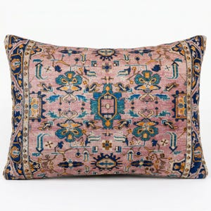May include: Rectangular decorative pillow with a vintage rug design. The pillow features a dusty rose background with a detailed pattern in shades of blue, teal, and gold. The edges are bordered with a dark blue and gold design.