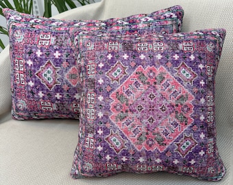 Op de afbeelding: Twee vierkante sierkussens met een levendig paars en roze geometrisch patroon. De kussens hebben een traditioneel tapijtontwerp met ingewikkelde details en een getextureerde uitstraling. Ze liggen op een neutraal gekleurde bank.