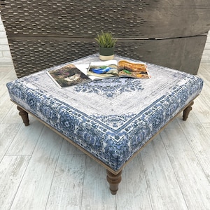 Pode incluir: Pufe quadrado com tampo de tecido estampado azul e cinza e pernas de madeira. O tecido tem um design detalhado e ornamentado. Revistas e uma pequena planta em vaso estão em cima do pufe.