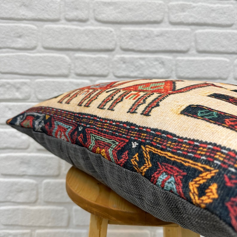 May include: Decorative pillow with a cream-colored background and a patterned design in red, black, and teal. The pillow has a dark gray border and is resting on a wooden stool. The pillow's design has a geometric pattern.