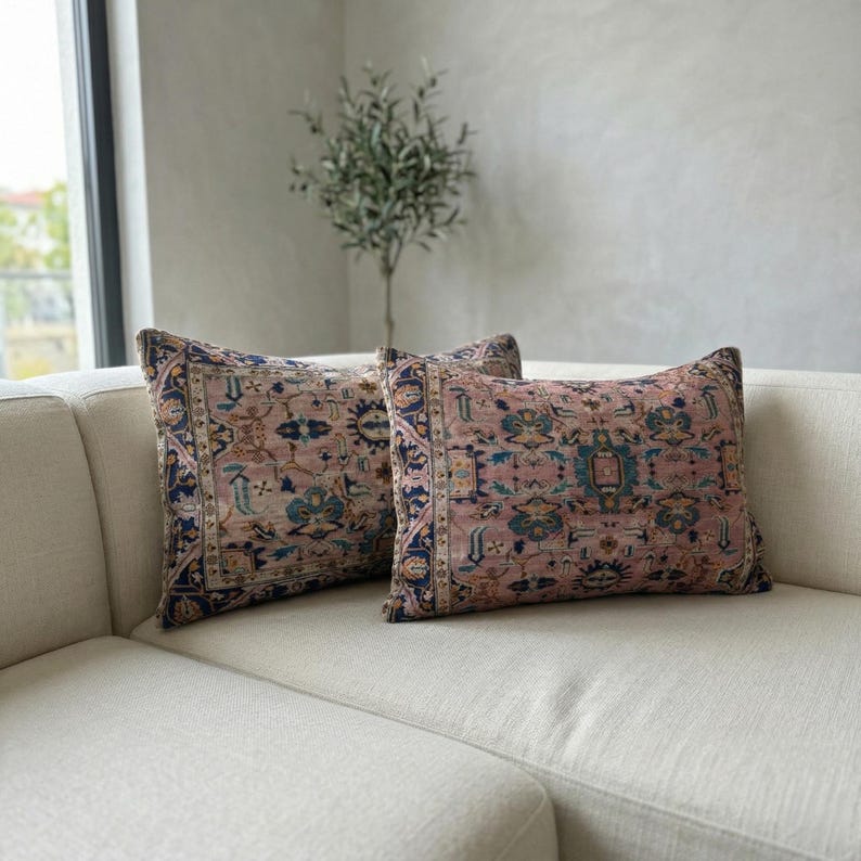May include: Two decorative throw pillows with a vintage rug design in shades of pink, blue, and cream. The pillows are rectangular and placed on a cream-colored sofa. The design features intricate patterns and a border.