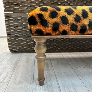 May include: A small bench with a leopard print seat cushion in shades of orange and black. The bench has a wooden frame and legs. The legs are light brown and have a turned design. The background is a woven basket.