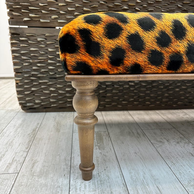 May include: A small bench with a leopard print seat cushion in shades of orange and black. The bench has a wooden frame and legs. The legs are light brown and have a turned design. The background is a woven basket.