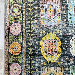May include: A close-up of a dark gray rug with a colorful geometric pattern. The rug features a variety of shapes and colors, including yellow, pink, blue, and green. The rug has a fringe border.