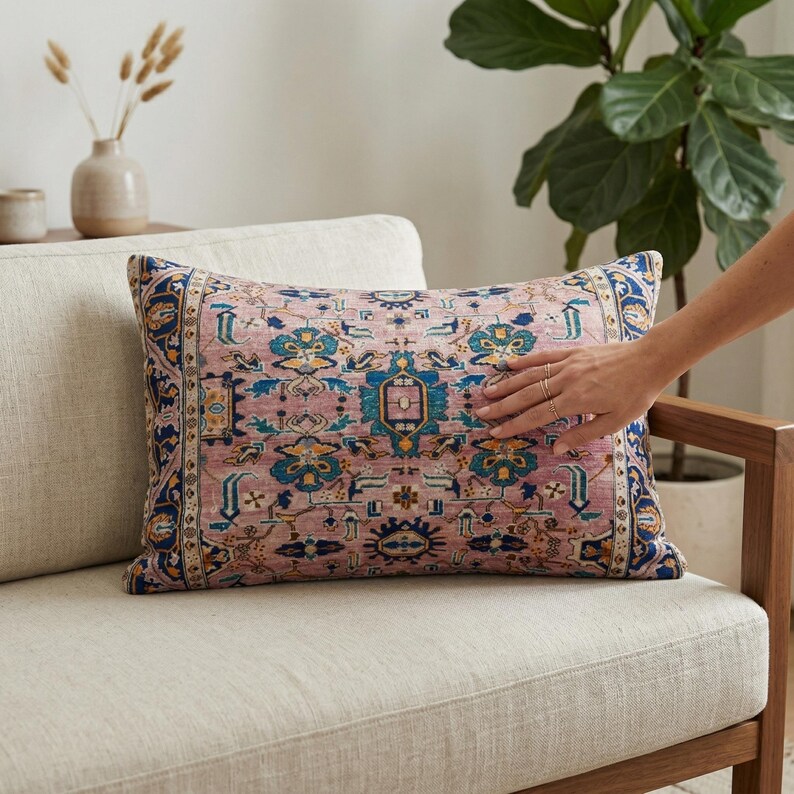 May include: Rectangular decorative pillow with a pink background and a detailed blue, yellow, and teal pattern. The pillow is placed on a cream-colored sofa. The design resembles a traditional rug.